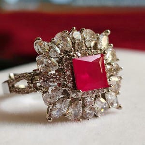 May include: A silver-toned ring featuring a central square-cut vibrant red gemstone surrounded by a halo of numerous clear, pear-shaped and round-cut stones. The band has a split design with small clear stones embedded along the metal surface.