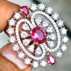 Könnte beinhalten: Eine kreisförmige Brosche mit einem großen, oval geschliffenen, leuchtend pinken Mittelstein, umgeben von einem wirbelnden Muster aus klaren weißen Steinen. Das Design umfasst mehrere tropfenförmige pinke Steine und zahlreiche runde klare Steine.