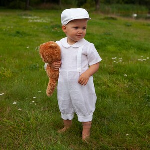 May include: A white short-sleeved romper with suspender-style straps and a rounded collar, paired with a matching white flat cap. The outfit is made of a light-colored fabric and features simple button details on the front straps and leg cuffs.