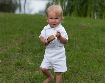 May include: A young boy wearing a white short-sleeved button-down shirt and white shorts stands in a grassy field. He is holding a small, brown, patterned object in his hand.
