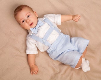 May include: A baby boy wearing a white linen shirt with a light blue bow tie and matching light blue shorts with suspenders. The baby is lying on a light brown fabric background.
