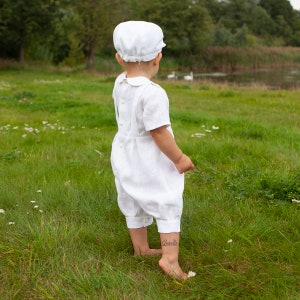 May include: A white linen romper featuring short sleeves, a Peter Pan collar, and a button-down back closure, paired with a matching white flat cap. The garment is designed with a pleated waist and cuffed leg openings, providing a classic look.