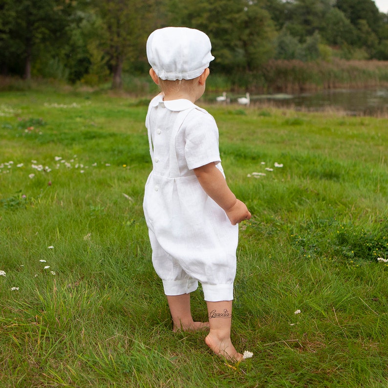 May include: A white linen romper featuring short sleeves, a Peter Pan collar, and a button-down back closure, paired with a matching white flat cap. The garment is designed with a pleated waist and cuffed leg openings, providing a classic look.
