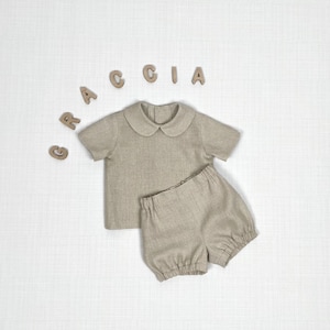 Puede incluir: Un conjunto beige de camisa de manga corta con cuello redondo y pantalones cortos con cintura elástica. Las letras de madera que forman la palabra GRACCIA están dispuestas en arco sobre la ropa, colocada sobre una superficie blanca.