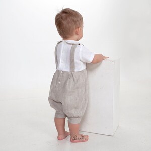 Puede incluir: Un conjunto para ni&ntilde;o peque&ntilde;o que consiste en una camisa blanca de manga corta con cuello redondo y un pantal&oacute;n corto beige con tirantes. La camisa tiene botones en la espalda y el pantal&oacute;n cuenta con cierre de bot&oacute;n en la cintura y pliegues.