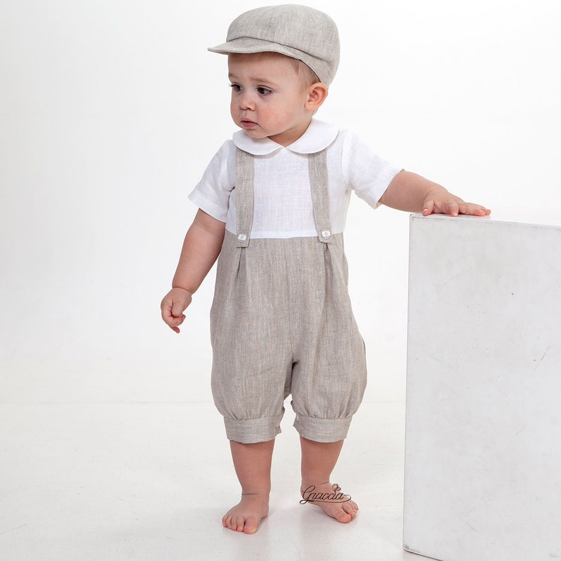 Puede incluir: Una gorra plana de color beige, una camisa blanca de manga corta con cuello redondo y un pantal&oacute;n corto con tirantes de color beige con botones en la parte delantera. El conjunto presenta una tela texturizada y un corte cl&aacute;sico.