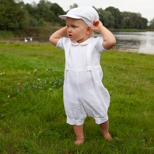 May include: A white short-sleeved romper with a Peter Pan collar and attached suspender straps featuring two buttons on the front. The outfit includes a matching white flat cap with a short brim, creating a coordinated two-piece set.