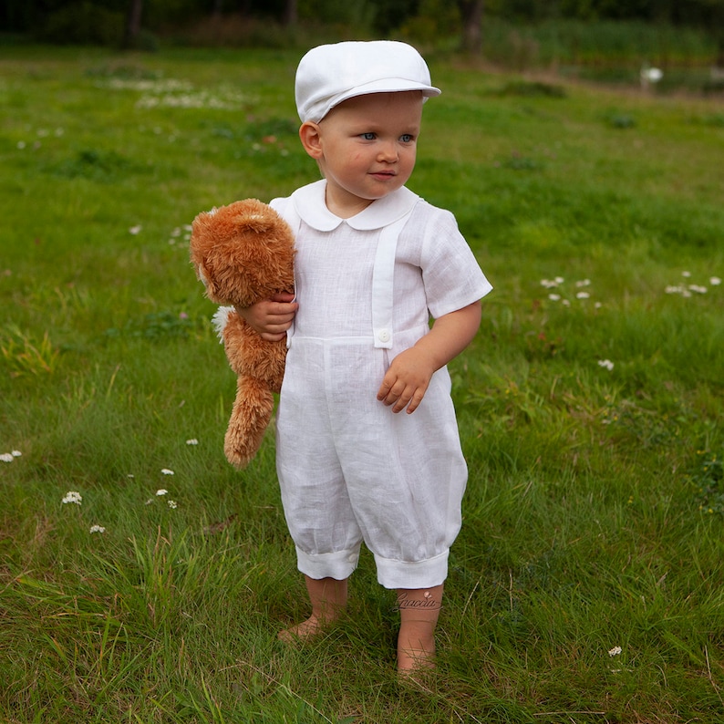 May include: A white short-sleeved romper with suspender-style straps and a rounded collar, paired with a matching white flat cap. The outfit is made of a light-colored fabric and features simple button details on the front straps and leg cuffs.