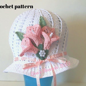 May include: White crochet hat with a pink flower embellishment and a pink ribbon. The hat has a wide brim and is decorated with a cluster of pink flowers and green leaves. The text "Bea's Bonnets 2015" is visible on the ribbon.