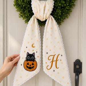 May include: A cream-colored fabric ribbon with a scalloped white edge, featuring a repeating pattern of small orange stars. One end displays an embroidered black cat inside an orange jack-o'-lantern with a crescent moon, and the other features a large letter H.