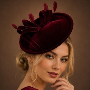 Pode incluir: Um fascinator de veludo bordô profundo com uma base redonda em forma de disco, adornado com laços de veludo estruturados e três penas delicadas a condizer que se estendem para cima a partir do centro do arranjo decorativo.