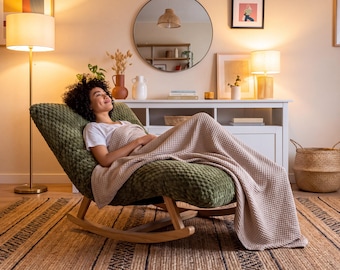 May include: A dark olive green textured rocking chair with a curved wooden base. A person is reclining in the chair, covered by a light beige waffle-knit blanket while wearing a plain white t-shirt. The chair features a soft, dimpled fabric surface.