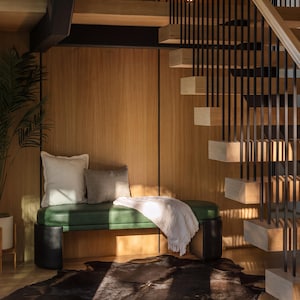 May include: A green upholstered bench with black cylindrical legs, topped with one large white square pillow and one smaller gray rectangular pillow, draped with a white throw blanket, positioned under a wooden staircase with black metal railings.