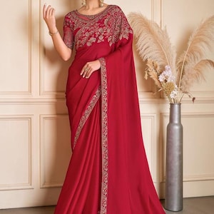 Può includere: Un sari rosso intenso caratterizzato da intricati ricami dorati lungo i bordi e il corpetto. L'indumento include una blusa trasparente a maniche corte con fitti motivi floreali e una gonna lunga e fluida con un bordo decorativo dorato.