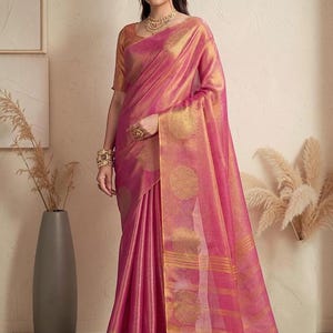 Könnte beinhalten: Ein leuchtend pinkfarbener Sari mit komplexen goldenen Kreismustern und horizontalen goldenen Streifen entlang des Randes. Das Kleidungsstück umfasst eine passende kurzärmelige Bluse mit metallischem Goldfinish sowie eine goldene Kette und Armreifen.