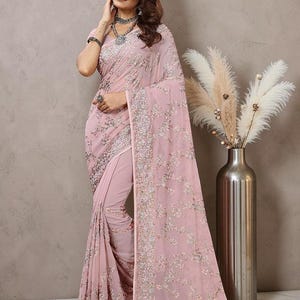 May include: A light pink saree featuring intricate silver embroidery and sequin detailing throughout the fabric. The garment includes a wide, heavily embellished border with floral patterns and a matching blouse with short sleeves and a round neckline.