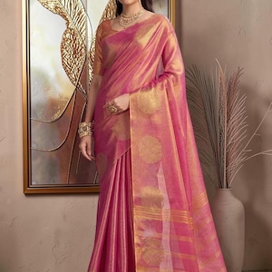 Könnte beinhalten: Ein leuchtend pinkfarbener Sari mit komplizierten goldenen kreisförmigen Motiven und horizontalen goldenen Streifen entlang des Randes. Das Kleidungsstück umfasst eine passende kurzärmelige Bluse mit leichtem Glanz, die elegant drapiert ist.