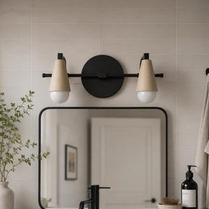 May include: A two-light wall sconce featuring a matte black circular backplate and horizontal bar, paired with two conical tan shades and spherical white glass bulbs. The fixture is mounted above a rectangular mirror with rounded corners.