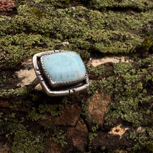 May include: A silver ring featuring a large, square-shaped, light blue Larimar gemstone with a smooth, polished surface. The stone is set in a thick, textured silver bezel with decorative notches along the edges of the metal frame.