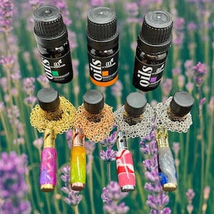 May include: Five small glass bottles with black caps and gold labels that say "oils" and "art naturals" are arranged in a row. The bottles are filled with essential oils.  Each bottle is attached to a gold or silver chain with a decorative pendant. The pendants are made of wood and have different colours and patterns. The bottles and pendants are on a background of purple lavender flowers.