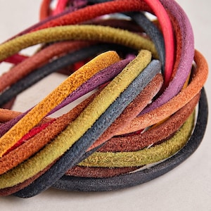 Può includere: Una collezione di cordini in pelle scamosciata multicolore avvolti insieme. I cordini rotondi presentano una superficie morbida e testurizzata in varie tonalità vivaci, tra cui giallo senape, verde oliva, viola intenso, rosso, arancione e nero.