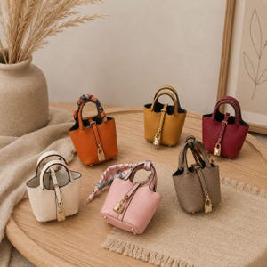 May include: Six miniature bucket bags in orange, yellow, burgundy, white, pink, and taupe. Each bag features a textured surface, two top handles, a central vertical strap with a gold-toned padlock closure, and four small metal feet at the base.