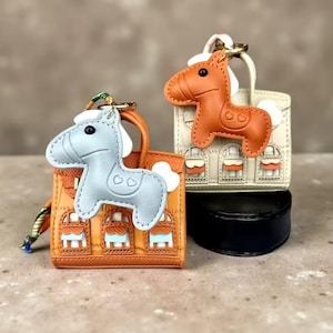 Op de afbeelding: Twee miniatuur handtasbedels met een lichtblauw paard en een oranje paard. Elk paard is bevestigd aan een kleine gestructureerde tas met een gevelontwerp. De oranje tas heeft lichtblauwe accenten, terwijl de crèmekleurige tas oranje accenten heeft.
