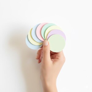 Könnte beinhalten: Eine Sammlung von acht kreisförmigen Papierdiscs in verschiedenen Pastellfarben wie Hellblau, Blassgelb, Zartrosa, Mintgrün und Lavendel, die fächerförmig angeordnet und als Stapel vor einem schlichten weißen Hintergrund gehalten werden.
