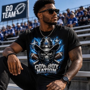 Op de afbeelding: Een zwart t-shirt met een afbeelding van een schedel met een cowboyhoed en een footballhelm, geflankeerd door twee footballs en twee sterren, omringd door blauwe bliksem, met de tekst COWBOY NATION in dikke blauwe en witte letters.