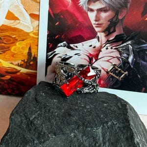 May include: A silver-toned metal ring featuring an intricate, textured band with a prominent, rectangular, faceted red gemstone set in the center. The ring has a dark, oxidized finish and an open-back design, showcasing detailed metalwork.