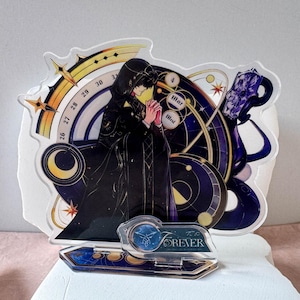 May include: A clear acrylic standee featuring a hooded figure in a black robe with hands clasped in prayer. The background includes celestial motifs, a calendar dial with numbers 26-31, and the text "Mar" and "FOREVER" on a blue base.