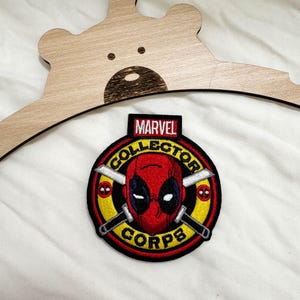 May include: A circular embroidered patch featuring the red Deadpool mask in the center with two crossed silver swords. The design includes a yellow border with the black text COLLECTOR CORPS and a red tab at the top with the white text MARVEL.
