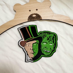May include: An embroidered patch featuring a split-face character wearing a tall black top hat with a brown band, transitioning from a natural skin tone to a bright green face, positioned next to a second green, snarling, monstrous face.