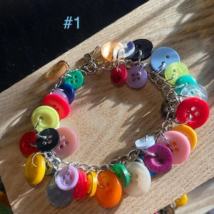 May include: A charm bracelet featuring a silver-toned metal chain adorned with numerous round buttons in various colors including red, blue, yellow, green, orange, purple, pink, black, and white. The text #1 is visible in the upper left corner.