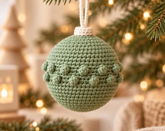 May include: A sage green crocheted sphere ornament featuring a textured band of small raised bobble stitches around the center. The ornament is topped with a cream-colored crocheted cap and a matching loop for hanging from a tree branch.