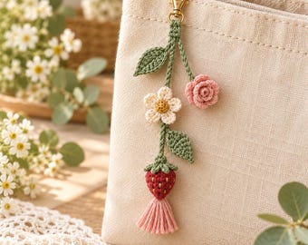 May include: A crochet bag charm featuring a red strawberry with a pink tassel, a white flower with a yellow centre, and a small pink rose, all connected by green stems with leaves to a gold-toned metal lobster clasp attached to a beige canvas bag.