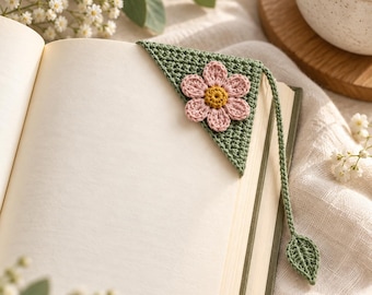 May include: A triangular crochet corner bookmark in sage green featuring a pink flower with a yellow centre attached to the top. A long, thin green crochet cord extends from the corner, ending in a small, detailed green crochet leaf shape.