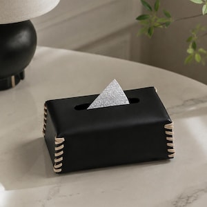 Puede incluir: Una caja rectangular negra para pañuelos con acabado liso y detalles de cordones en color beige claro a lo largo de los bordes verticales. La parte superior tiene una ranura horizontal centrada con un pañuelo blanco asomando.