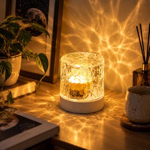 May include: A cylindrical table lamp with a textured, clear glass shade that emits a warm, golden light, creating intricate, rippling patterns on the surrounding surface. The lamp features a solid white base and a compact, modern design.