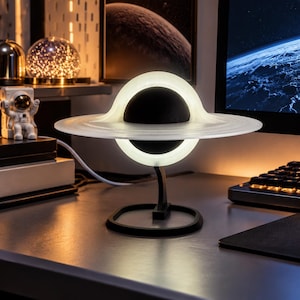 Könnte beinhalten: Eine schwarze kugelförmige Lampe mit einer leuchtend weißen Ringstruktur, die einem Planeten mit Ringen ähnelt. Die Lampe wird von einem schwarzen Ständer mit rundem Fuß getragen und leuchtet, wodurch ein sanftes Licht auf den Schreibtisch fällt.