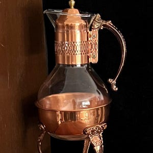 May include: A clear glass carafe featuring a polished copper base and a matching copper lid with a decorative lattice band. The carafe includes an ornate, curved copper handle and rests on a three-legged copper stand with intricate floral detailing.
