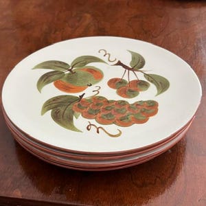 May include: A stack of three round ceramic plates with a white base and a painted fruit motif. The design features a cluster of orange grapes, a single orange peach, and a small bunch of red cherries, all accented with green leaves.