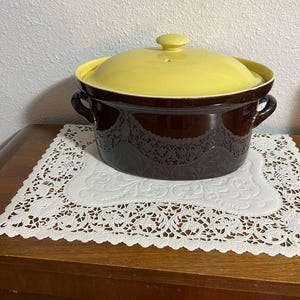 May include: A dark brown ceramic casserole dish with a glossy finish and a matching light yellow lid. The pot features two side handles and a decorative embossed pattern around the exterior base. The lid has a small central knob handle.