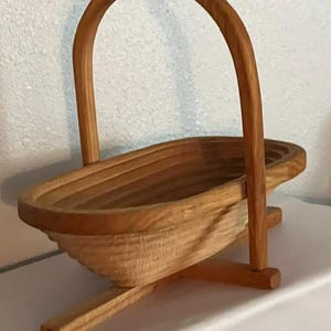 May include: A light brown wooden basket with a smooth, curved handle and a ribbed, oval-shaped body. The basket is supported by a cross-shaped wooden base that extends outward, providing stability for the structure while maintaining a natural wood grain.