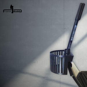 Könnte beinhalten: Ein schwarzer, 3D-gedruckter Getränkehalter mit halbkreisförmigem Lamellendesign und einer zylindrischen Öffnung, in der ein schwarzer Stift steckt. Das Frame Buddy-Logo ist oben links zu sehen. Das Zubehör ist an einer Kante befestigt.