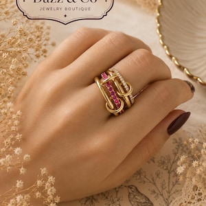 Puede incluir: Un anillo de oro de varias bandas que presenta una banda con pequeñas piedras rosas y otra banda con piedras blancas transparentes. El anillo tiene un diseño de eslabones entrelazados. El texto Dazz & Co Jewelry Boutique y Timeless Style. Elevated Details. es visible.