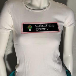 Può includere: Una t-shirt bianca a maniche corte con una grafica rettangolare nera sul petto. La grafica contiene un piccolo albero verde con frutti rossi e il testo bianco "organically grown", il tutto contornato da un sottile bordo rosso e bianco.