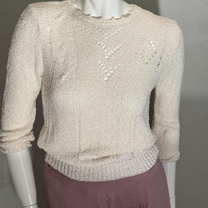 May include: A cream-colored, textured knit sweater with three-quarter length sleeves, a round neckline, and subtle openwork detailing on the chest, paired with mauve-colored trousers featuring a pleated waistline.