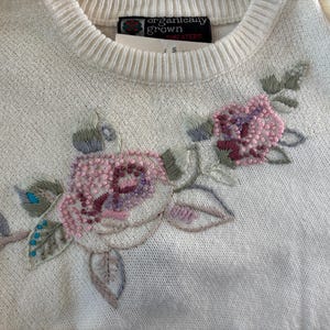 May include: A cream-colored knit sweater featuring an embroidered floral design with pink, purple, and sage green thread and bead accents. The interior neck label reads Organically Grown Sweaters. The sweater has a ribbed crew neckline.
