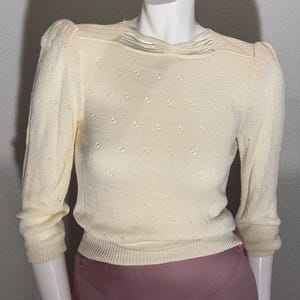 May include: A cream-colored knit sweater featuring three-quarter length sleeves, a round neckline with horizontal ribbed detailing, and a subtle pointelle pattern throughout the fabric. The sweater has a fitted ribbed hemline.